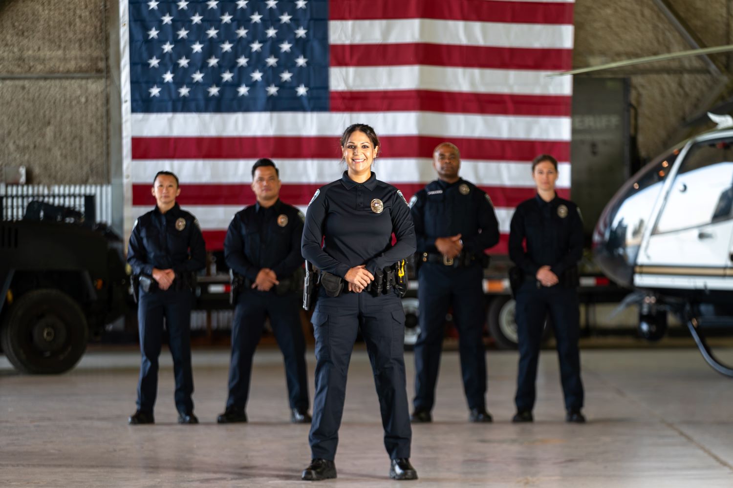Five law enforcement officers in navy duty uniforms standing in a hangar in front of an American flag, with an armored vehicle and a law enforcement helicopter